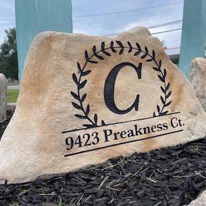 May include: A light brown stone with a black wreath and the letter "C" in the center. The stone also has the address "9423 Preakness Ct." engraved on it.