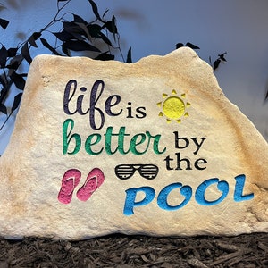 May include: A painted rock with the text "life is better by the pool" in black, green, blue, and yellow. The rock also features a sun, sunglasses, and flip-flops.