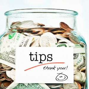 Op de afbeelding: Een glazen pot gevuld met Amerikaanse valuta, waaronder dollarbiljetten en munten, met een wit label waarop "tips thank you!" staat en een smiley.