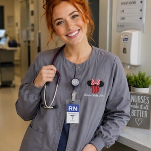 May include: A grey button-down scrub jacket with long sleeves and elastic cuffs. The jacket features an embroidered Minnie Mouse ear icon with a red polka dot bow above the name Theresa Vohn, RN. A stethoscope and an RN ID badge are worn.