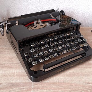 May include: A vintage black Corona typewriter with silver keys and a red ribbon. It's on a light wood surface, next to a patterned mug, a stack of books, and a decorative object. The typewriter is a classic writing tool.