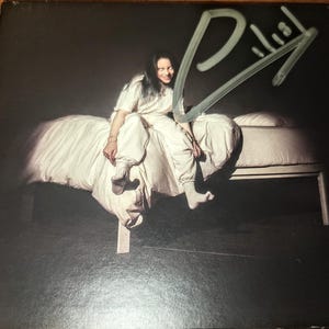 Puede incluir: Una portada de álbum firmada con una persona sentada en una cama. La persona lleva un atuendo blanco y tiene el pelo largo y oscuro. La portada del álbum es negra y tiene el título "When We Fall Asleep, Where Do We Go?" escrito en blanco.