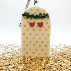 May include: A white ceramic ornament with a floral pattern and green and red glass accents. The ornament has a black ribbon for hanging.