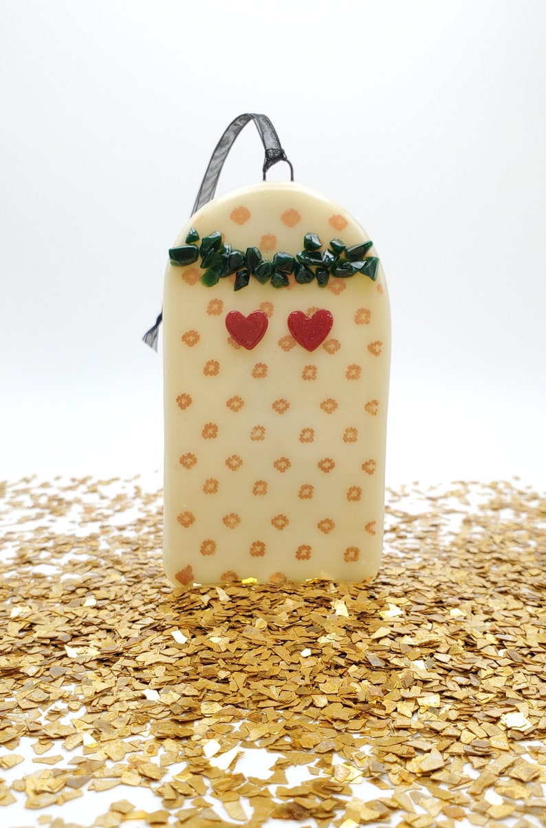 May include: A white ceramic ornament with a floral pattern and green and red glass accents. The ornament has a black ribbon for hanging.