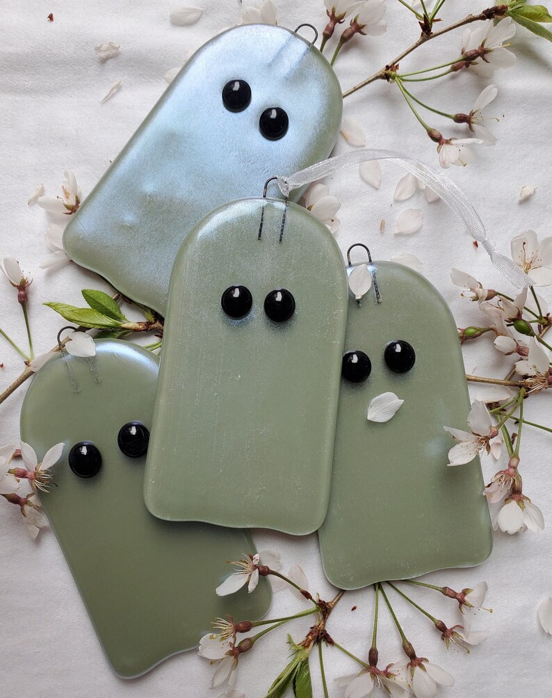 May include: Four glass ghost ornaments, two are green and two are iridescent blue. The ghosts have black eyes and are hanging from a white background.