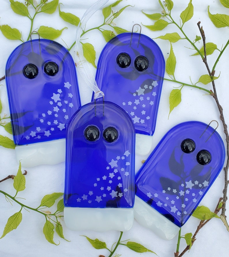 May include: Four blue glass ornaments with white stars and black eyes. The ornaments are shaped like ghosts and have a white bottom.