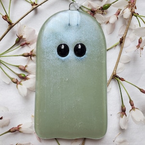 May include: A light green glass ghost ornament with black eyes. The ornament is hanging from a white ribbon and is surrounded by white blossoms.