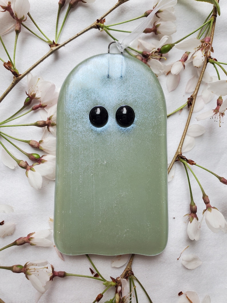 May include: A light green glass ghost ornament with black eyes. The ornament is hanging from a white ribbon and is surrounded by white blossoms.