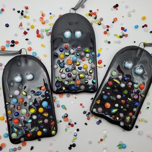 May include: Three black glass ghost ornaments with colorful glass beads embedded in the surface. The ghosts have white eyes and are hanging from black ribbon.