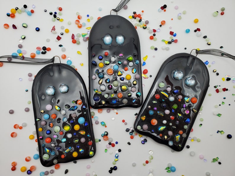 May include: Three black glass ghost ornaments with colorful glass beads embedded in the surface. The ghosts have white eyes and are hanging from black ribbon.