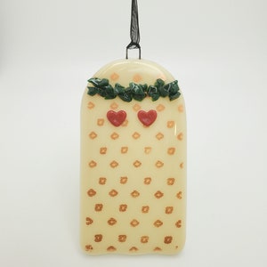 May include: A white glass ornament with a gold floral pattern and two red hearts. The ornament is topped with a green garland and has a black ribbon for hanging.