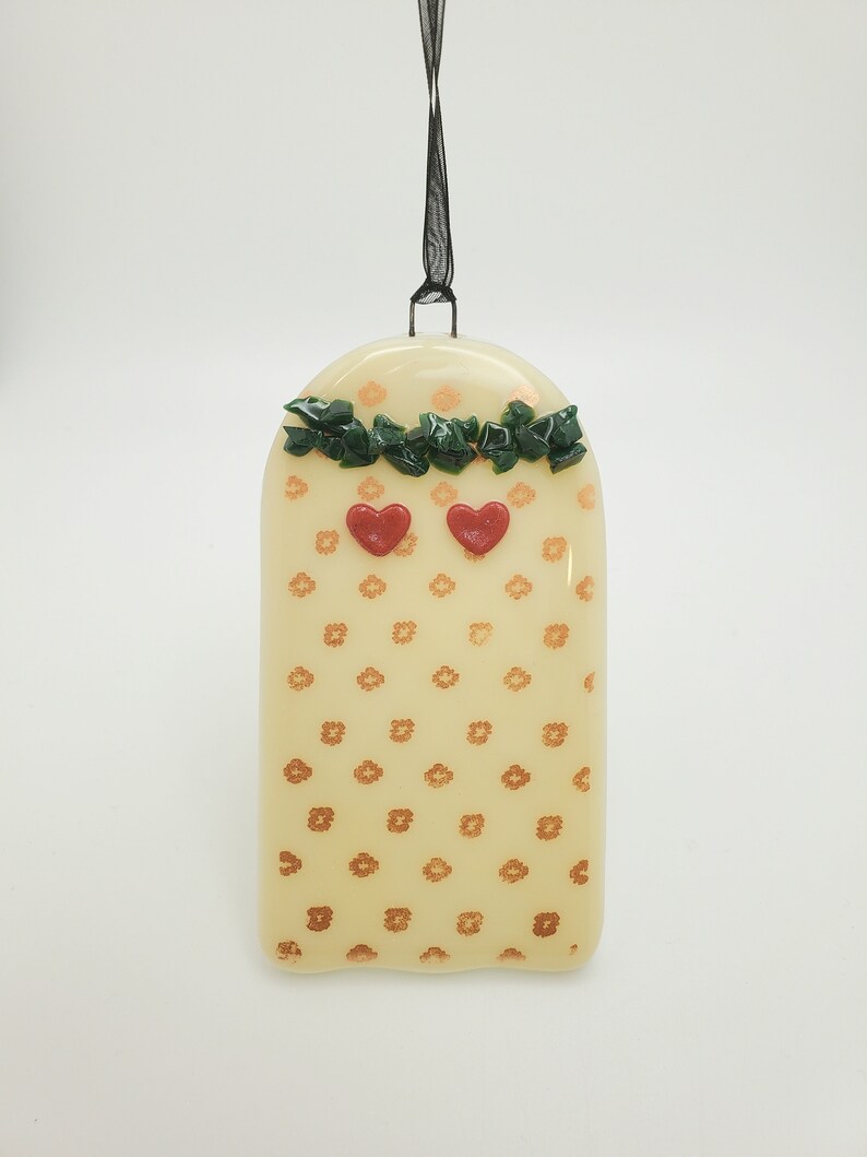 May include: A white glass ornament with a gold floral pattern and two red hearts. The ornament is topped with a green garland and has a black ribbon for hanging.