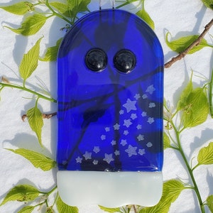 May include: A blue glass ghost ornament with white stars and black eyes. The ornament is hanging from a white ribbon.