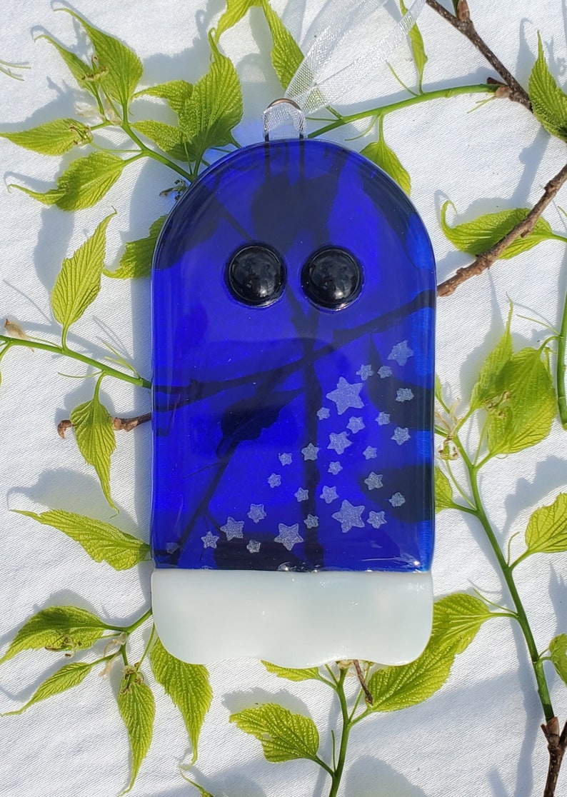 May include: A blue glass ghost ornament with white stars and black eyes. The ornament is hanging from a white ribbon.