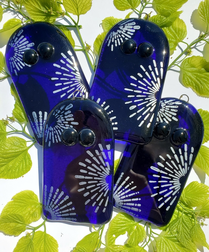May include: Four handmade glass ornaments in a deep blue hue, each featuring a stylized white burst design and two black glass bead eyes. The ornaments are arranged with green leaves in the background.