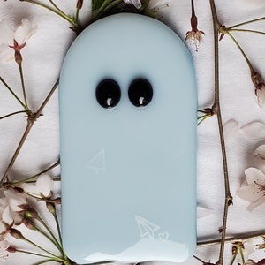 K&ouml;nnte beinhalten: Hellblaues, geisterf&ouml;rmiges Ornament mit zwei schwarzen Augen und einem Papierflugzeug-Design. Das Ornament h&auml;ngt an einem silbernen Band und ist von wei&szlig;en Bl&uuml;ten und Zweigen umgeben.
