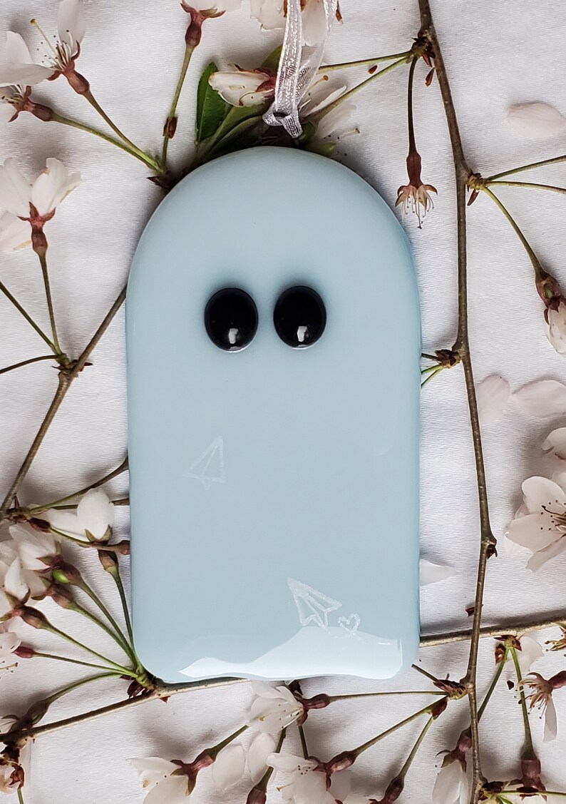 K&ouml;nnte beinhalten: Hellblaues, geisterf&ouml;rmiges Ornament mit zwei schwarzen Augen und einem Papierflugzeug-Design. Das Ornament h&auml;ngt an einem silbernen Band und ist von wei&szlig;en Bl&uuml;ten und Zweigen umgeben.