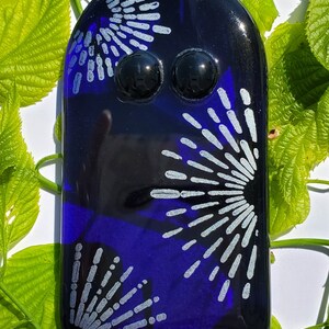 May include: A dark blue glass art piece with a rounded top and two black, spherical accents. The piece features a design of silver-colored radiating lines. A small metal loop is at the top for hanging. The background includes green leaves.