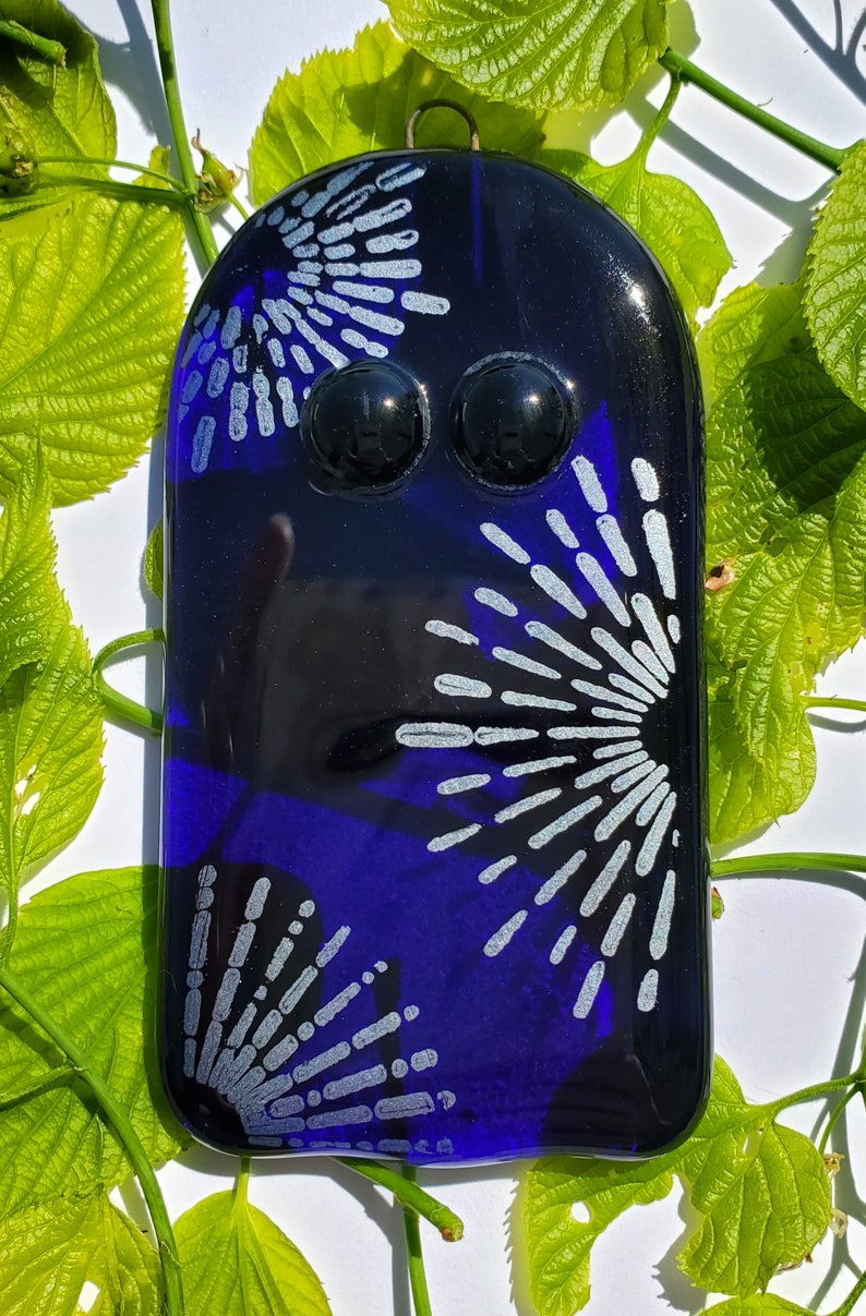 May include: A dark blue glass art piece with a rounded top and two black, spherical accents. The piece features a design of silver-colored radiating lines. A small metal loop is at the top for hanging. The background includes green leaves.