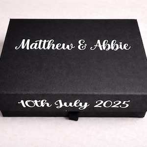 May include: A rectangular black gift box featuring the white printed text Matthew & Abbie on the lid and 10th July 2025 on the front side. The box has a smooth matte finish and a small black pull tab centered on the bottom edge.