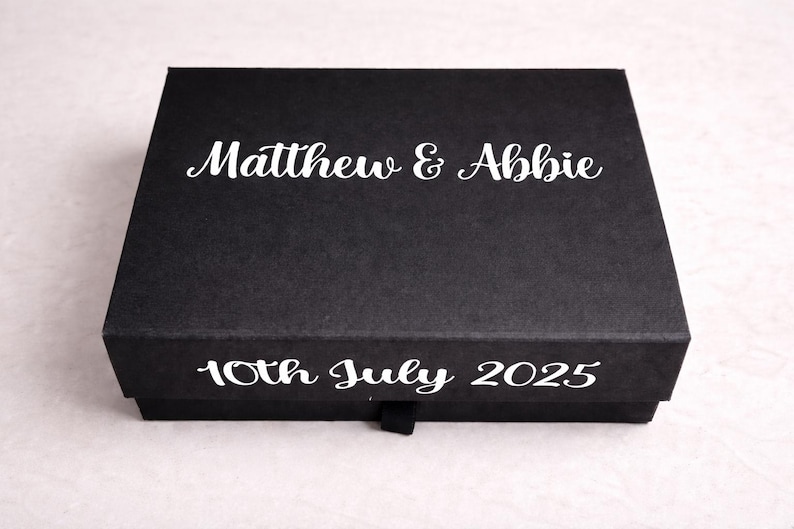 May include: A rectangular black gift box featuring the white printed text Matthew & Abbie on the lid and 10th July 2025 on the front side. The box has a smooth matte finish and a small black pull tab centered on the bottom edge.