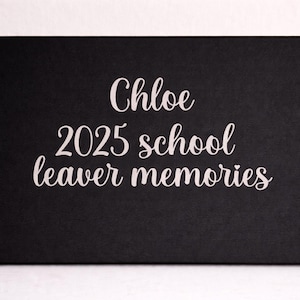 May include: A rectangular black box featuring white cursive text that reads Chloe 2025 school leaver memories. The box has a smooth, matte finish and a clean, minimalist design with the lettering centered on the front panel.