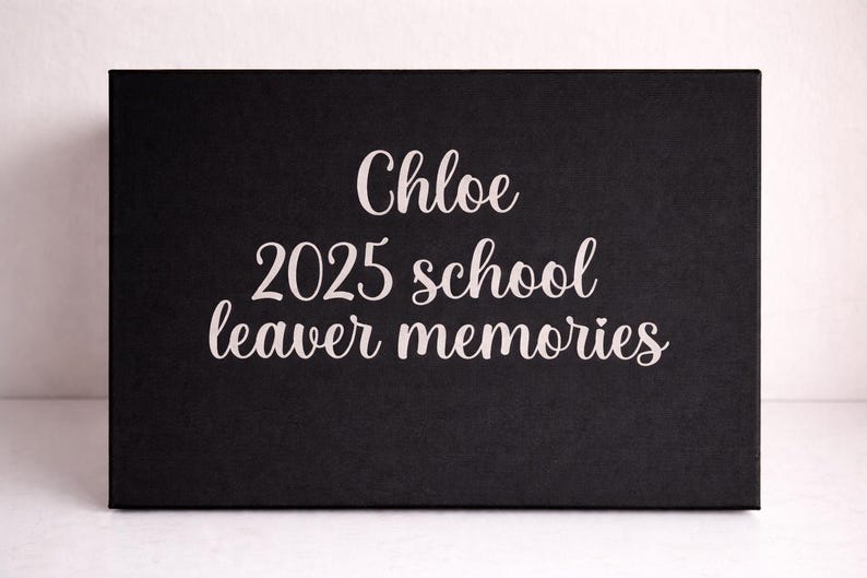 May include: A rectangular black box featuring white cursive text that reads Chloe 2025 school leaver memories. The box has a smooth, matte finish and a clean, minimalist design with the lettering centered on the front panel.