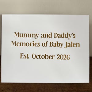 May include: A white rectangular sign featuring gold lettering that reads: Mummy and Daddy's Memories of Baby Jalen, Est. October 2026. The text is centered on the smooth surface, with a small heart icon above the word Memories.