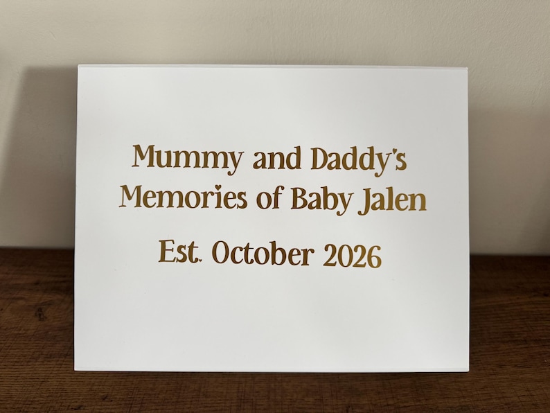 May include: A white rectangular sign featuring gold lettering that reads: Mummy and Daddy's Memories of Baby Jalen, Est. October 2026. The text is centered on the smooth surface, with a small heart icon above the word Memories.