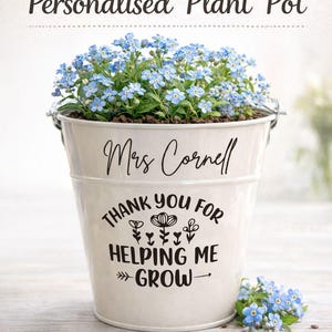 May include: A cream-coloured plant pot filled with blue flowers. The pot is inscribed with "Mrs Cornell" and "Thank you for helping me grow." A banner at the top reads "Teacher Gift." The pot has a metal handle.