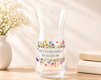 May include: A clear glass vase with a wide base and a narrow neck. The vase has a floral design with the text "MUM, IF YOU WERE A FLOWER, I'D PICK YOU EVERY TIME". The vase is sitting on a wooden surface.