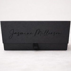 May include: A rectangular black gift box with a matte finish featuring the text Jasmine Millerson in a black script font on the front lid. The box includes a small black fabric pull tab centered at the bottom edge for opening the lid.
