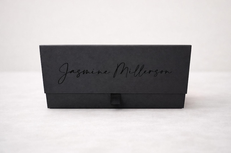 May include: A rectangular black gift box with a matte finish featuring the text Jasmine Millerson in a black script font on the front lid. The box includes a small black fabric pull tab centered at the bottom edge for opening the lid.
