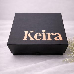 May include: A square black gift box with a textured finish. The lid features the name Keira printed in a metallic rose gold font, with a small heart icon replacing the dot above the letter i. The box has a simple, clean design.