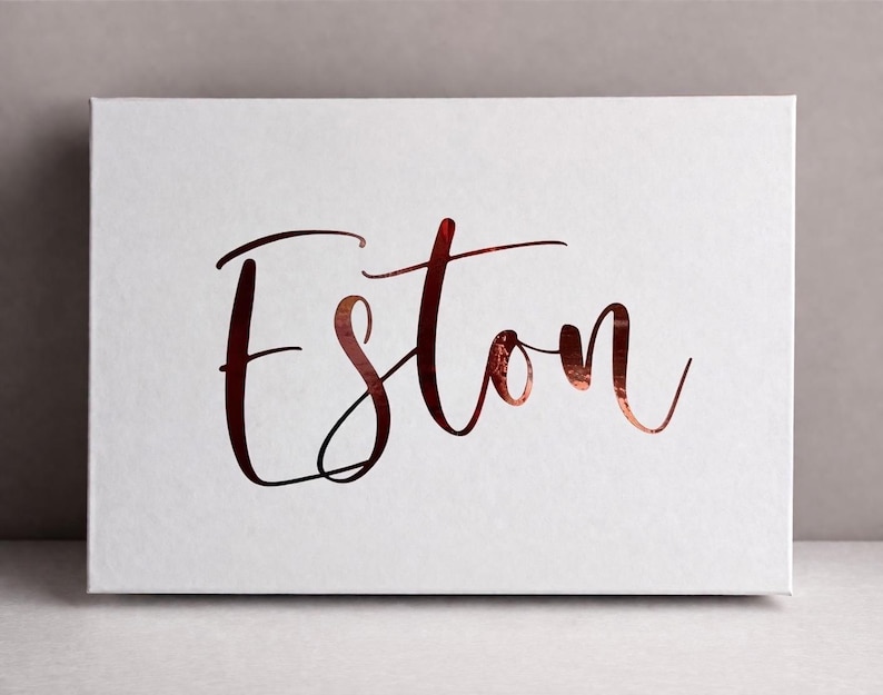 May include: A rectangular white canvas featuring the name Eston written in a metallic copper-colored script font. The lettering has a reflective, foil-like finish that contrasts with the matte white surface of the rectangular board.