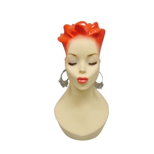 May include: A mannequin head with short, bright orange hair, closed eyes, and a neutral expression. The mannequin is wearing silver hoop earrings with dangling charms.