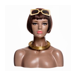 May include: A brown mannequin with short brown hair wearing a gold choker necklace and white sunglasses with gold accents.