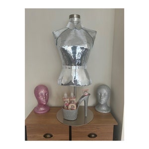 May include: A silver sequined mannequin torso stands above a decorative high-heeled shoe-shaped container holding beauty products. Two glittery head busts, one pink and one silver, flank the display on a wooden surface.