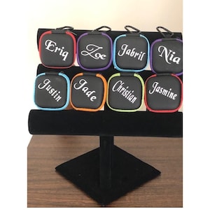 May include: Eight small black zippered pouches with personalized names in white lettering. The pouches are displayed on a black velvet stand. The pouches are in various colors: red, purple, blue, green, orange, and black.