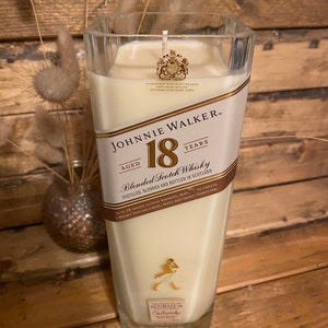 May include: A large, square glass candle with a cream-colored wax fill. The candle features a label with "JOHNNIE WALKER 18 YEARS" branding. The candle is set against a wooden background, with dried decorative plants in a vase.