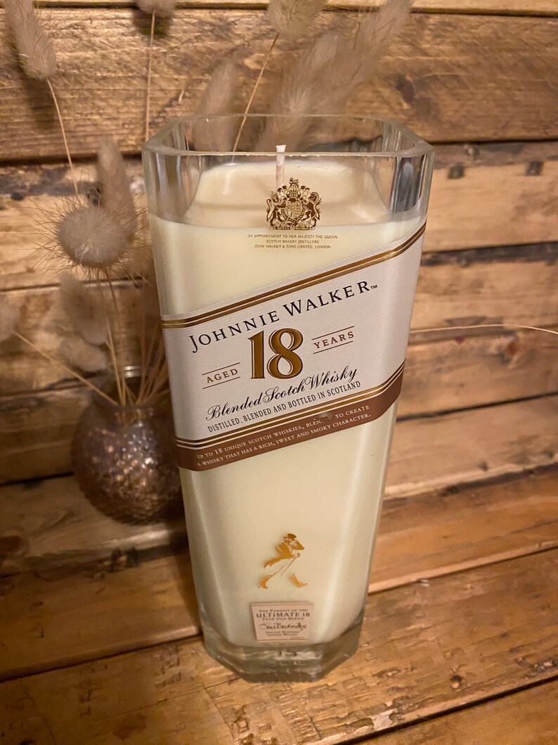 May include: A large, square glass candle with a cream-colored wax fill. The candle features a label with "JOHNNIE WALKER 18 YEARS" branding. The candle is set against a wooden background, with dried decorative plants in a vase.