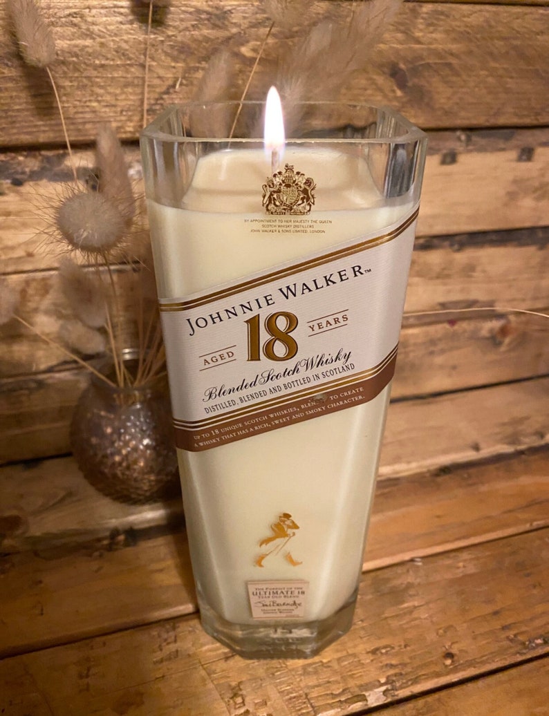 May include: A lit candle in a clear, square-shaped glass. The candle has a cream-colored wax and a label that reads "JOHNNIE WALKER 18 YEARS Blended Scotch Whisky." The label is white and gold with a logo of a man walking. The candle is on a wooden surface.
