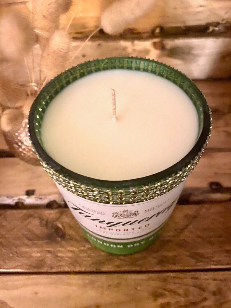 May include: A green glass candle jar with a gold glitter rim and a white candle inside. The jar is decorated with a label that says "Tanqueray" and "Imported London Dry Gin".