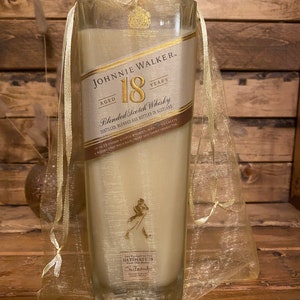 May include: A clear glass candle with a cream-colored wax fill, featuring a label that reads "JOHNNIE WALKER 18 YEARS." The candle is encased in a sheer gold organza bag, tied at the top with a matching ribbon. The label includes the text "Blended Scotch Whisky."
