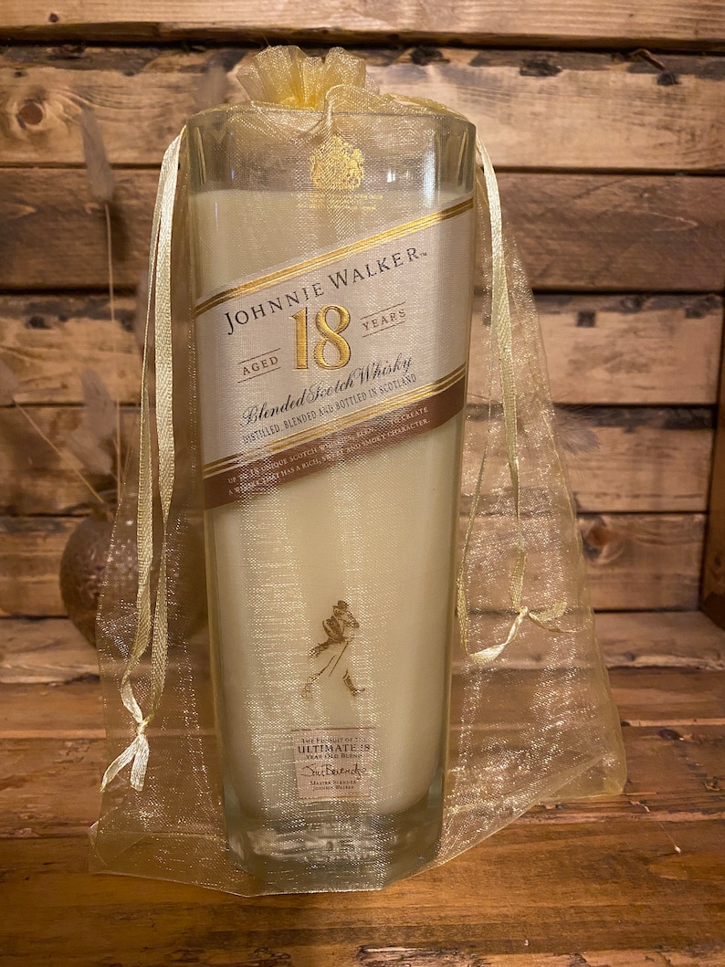 May include: A clear glass candle with a cream-colored wax fill, featuring a label that reads "JOHNNIE WALKER 18 YEARS." The candle is encased in a sheer gold organza bag, tied at the top with a matching ribbon. The label includes the text "Blended Scotch Whisky."