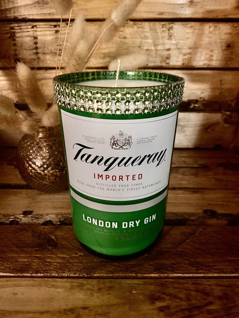 May include: A green glass candle jar with a gold and white glitter rim. The jar is labeled "Tanqueray Imported London Dry Gin" and features a crest with a lion and a unicorn.