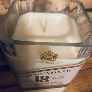 May include: A square glass candle holder filled with cream-colored wax. The candle has a single wick and is labeled "Johnnie Walker 18 Years Blended Scotch Whisky". The label includes a royal crest.