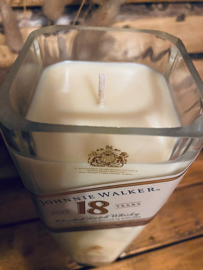 May include: A square glass candle holder filled with cream-colored wax. The candle has a single wick and is labeled "Johnnie Walker 18 Years Blended Scotch Whisky". The label includes a royal crest.