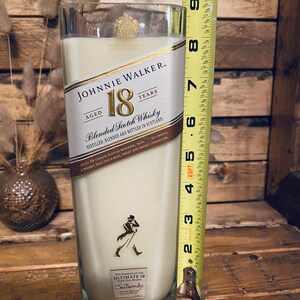 May include: A tall, rectangular candle in a repurposed Johnnie Walker 18 Years Aged Blended Scotch Whisky bottle. The candle is filled with a creamy wax and measures approximately 8 inches tall. The label is still attached, and the candle has a smoky character.