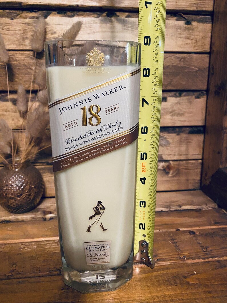 May include: A tall, rectangular candle in a repurposed Johnnie Walker 18 Years Aged Blended Scotch Whisky bottle. The candle is filled with a creamy wax and measures approximately 8 inches tall. The label is still attached, and the candle has a smoky character.